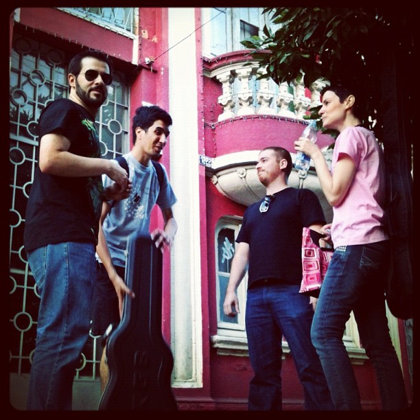 Band members beside a building facade under colored light.